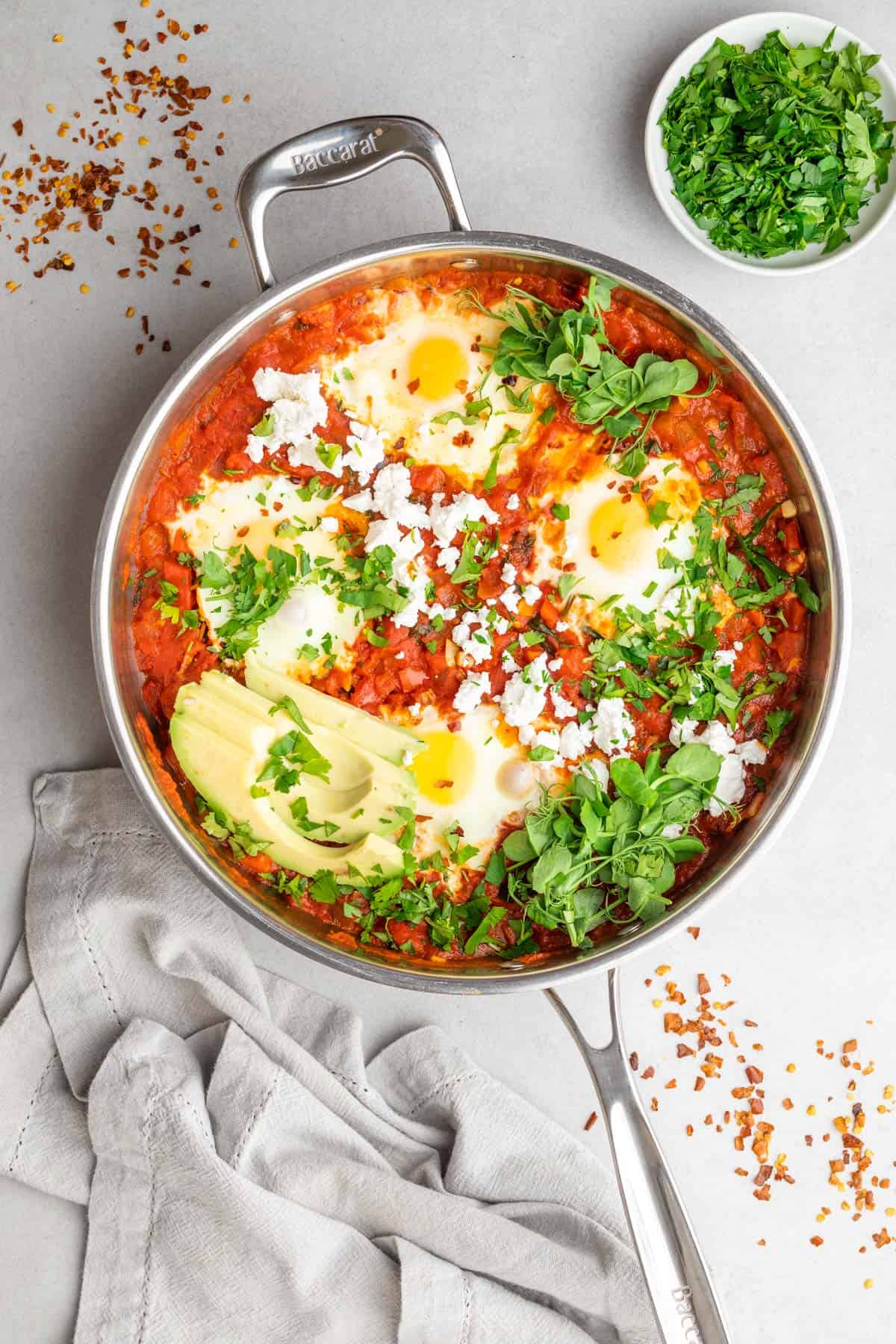 Shakshuka in a stainless steel pan with green herbs and garnishes