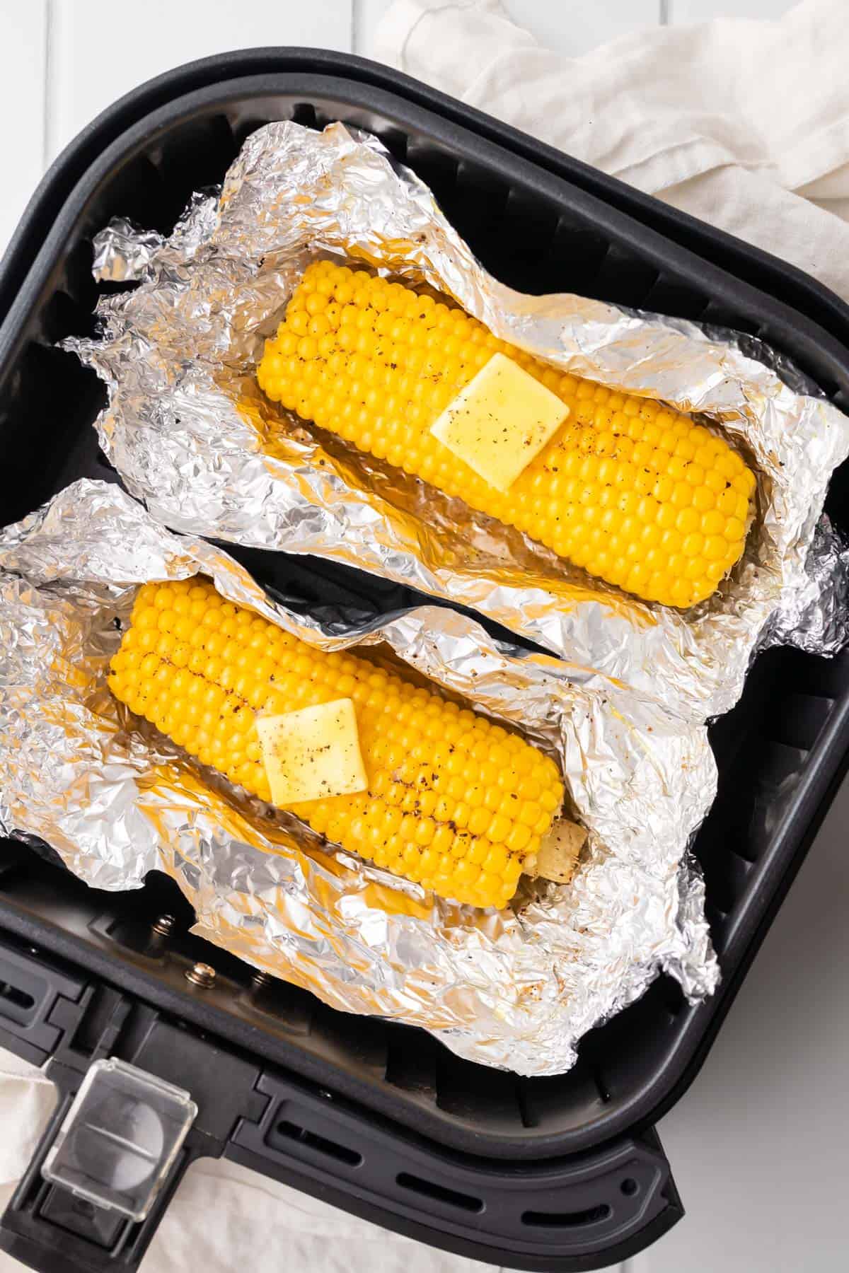 two corn cobs unwrapped in the air fryer basket garnished with butter and black pepper