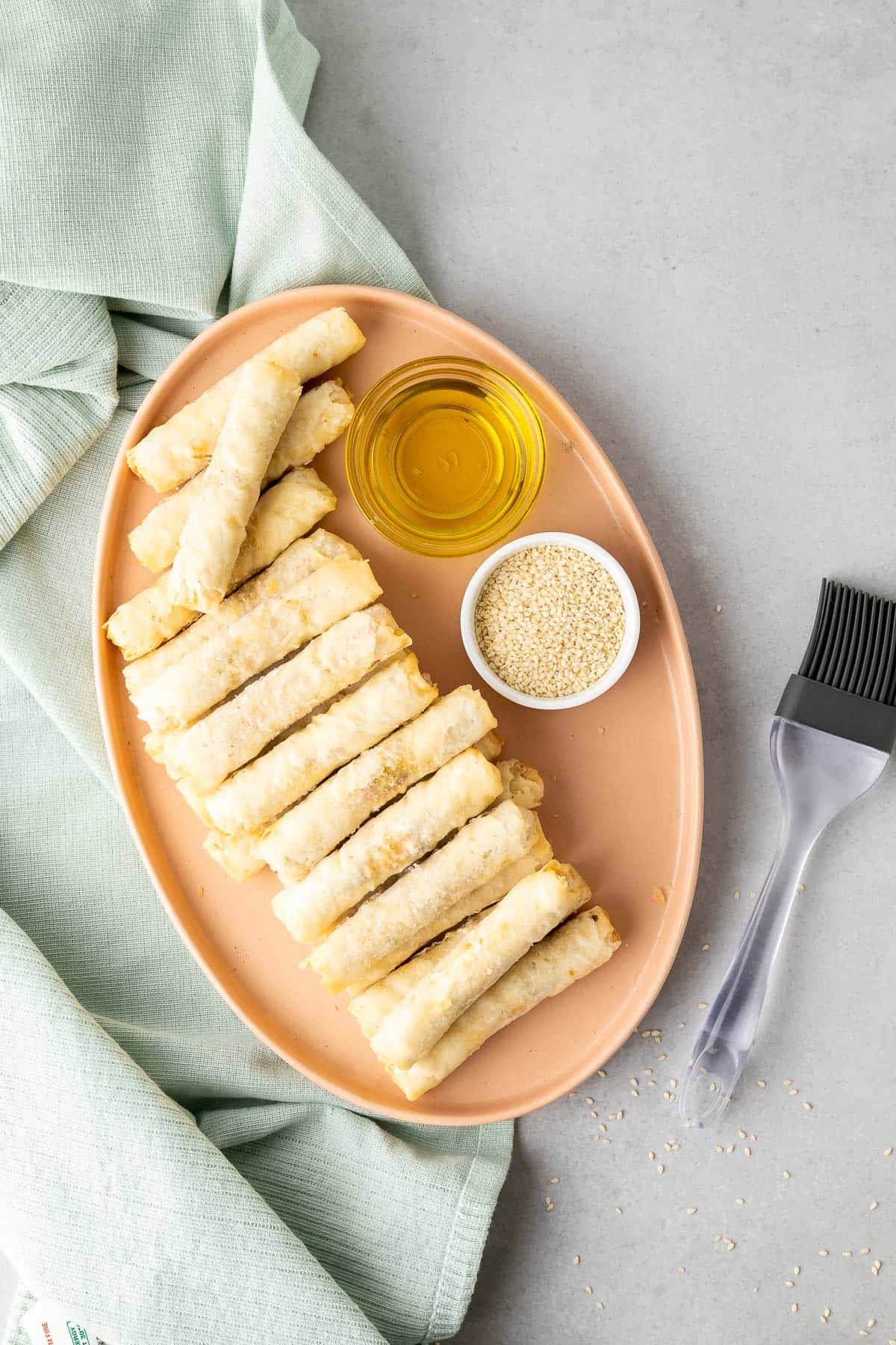 frozen egg rolls, oil and white sesame seeds on a platter with a silicone brush and green cloth on either side