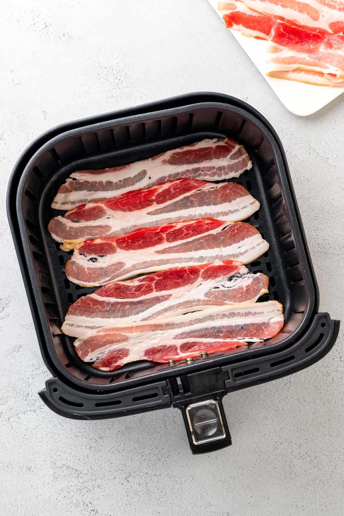 raw strips of bacon laid out in neat rows in the air fryer basket