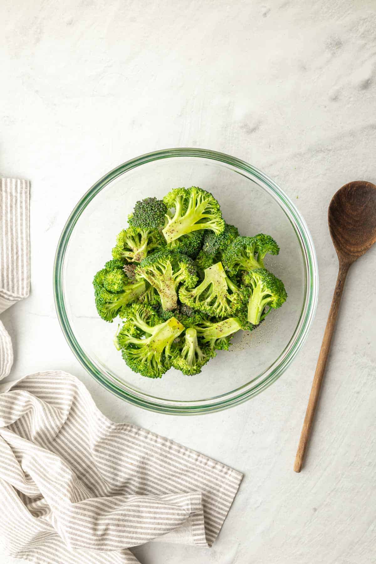 broccoli florets in a glass bowl with seasonings and a wooden spoon for mixing on one side