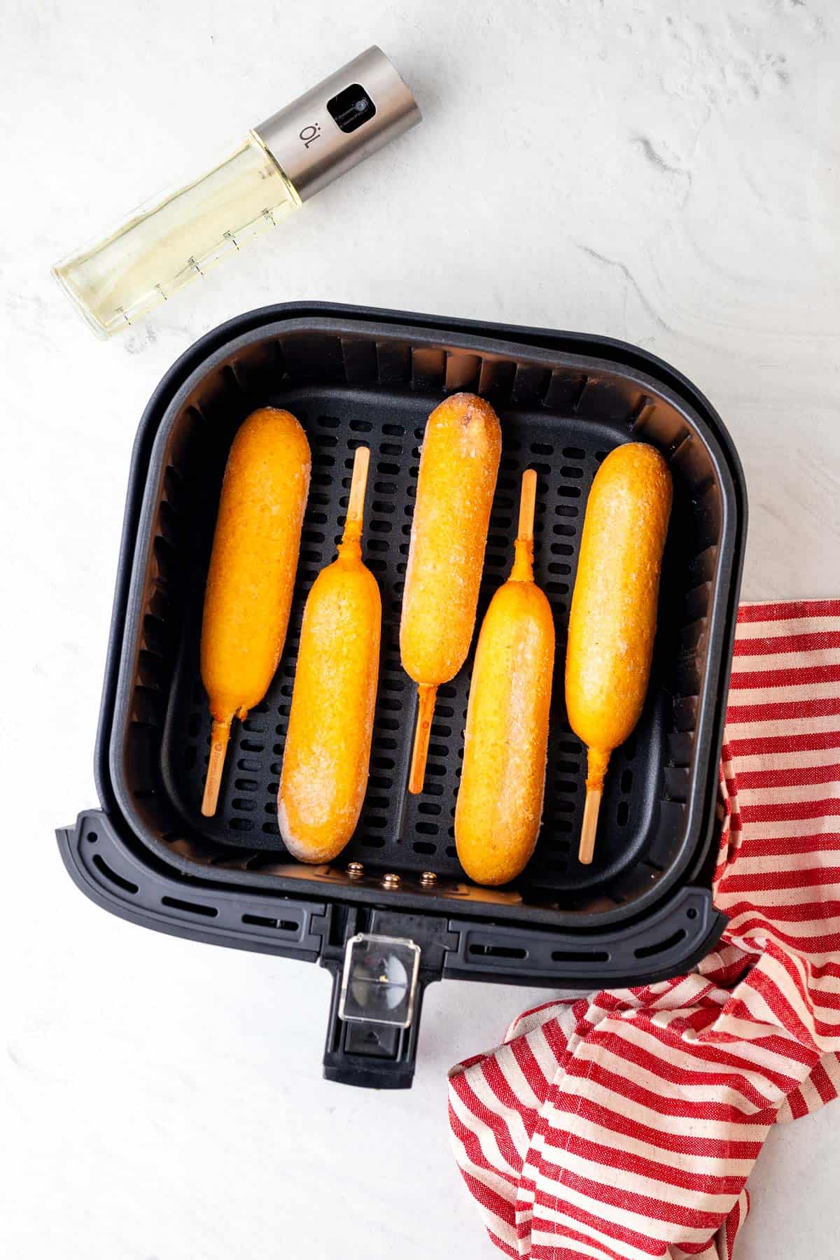 frozen corn dogs in the air fryer basket with an oil sprayer bottle to the one side of the air fryer basket and a red and white striped decorative cloth to the right