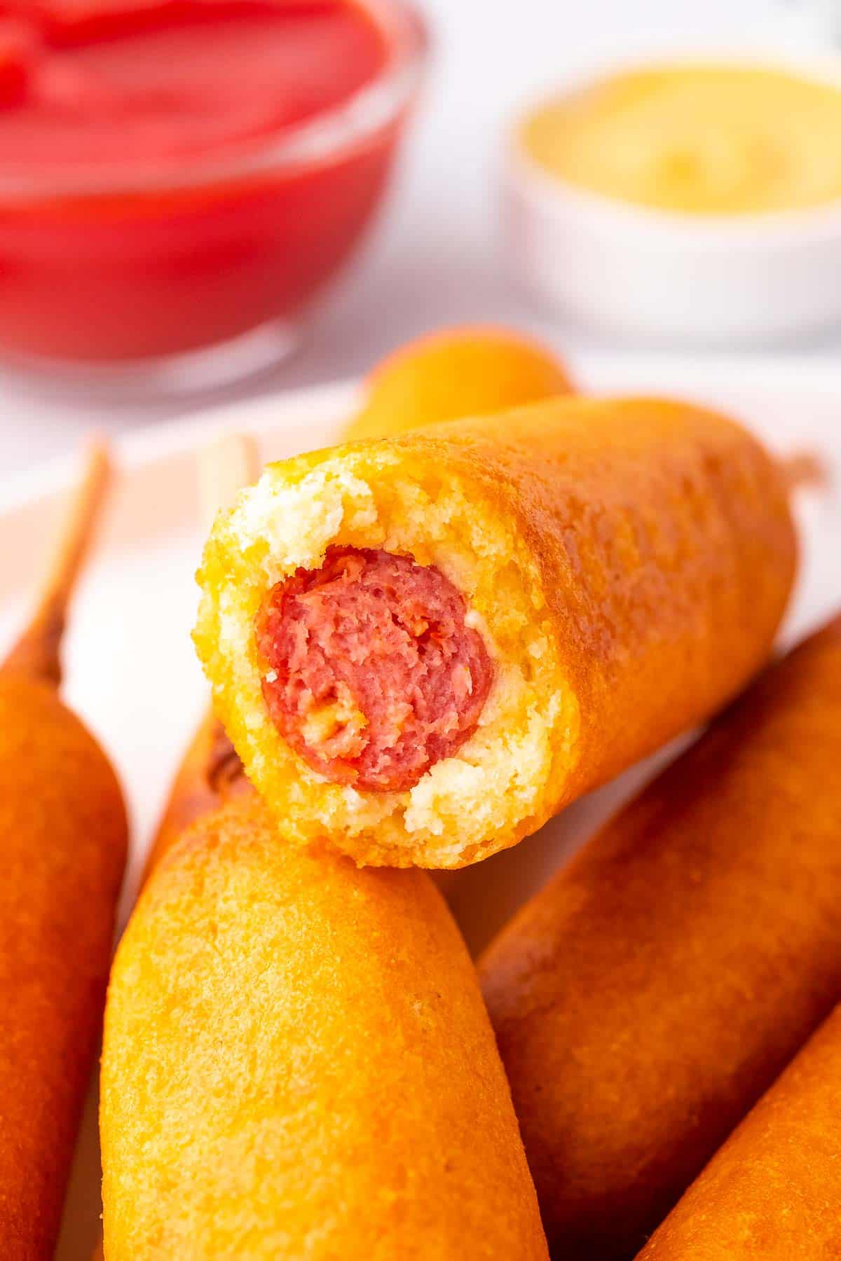 a close up of a corn dog on top of the corn dogs, the top corn dog has a bite taken out of it and there is ketchup and mustard in the background