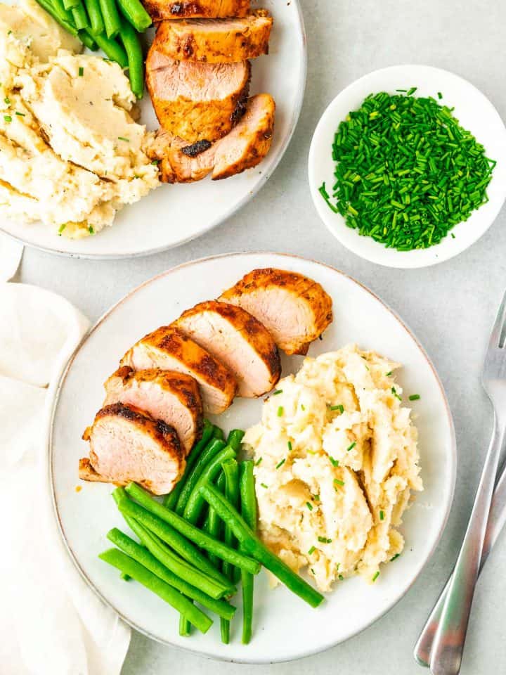 sliced pork tenderloin on a plate served with mashed potatoes and green beans, all garnished with fresh chopped chives