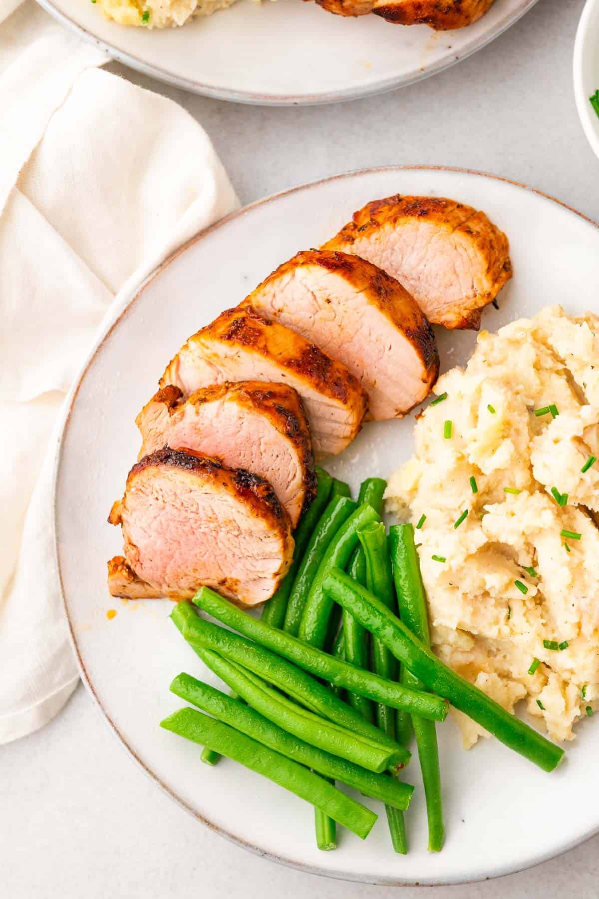 sliced cooked pork tenderloin on a plate served with mashed potatoes and green beans, all garnished with chopped chives