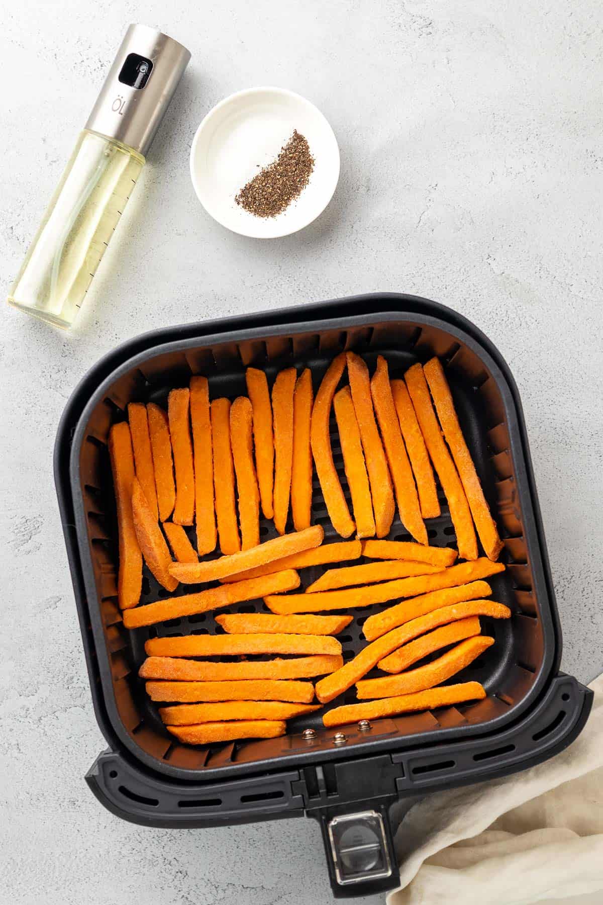 sweet potato fries in the air fryer basket with salt and pepper and an oil sprayer around the basket