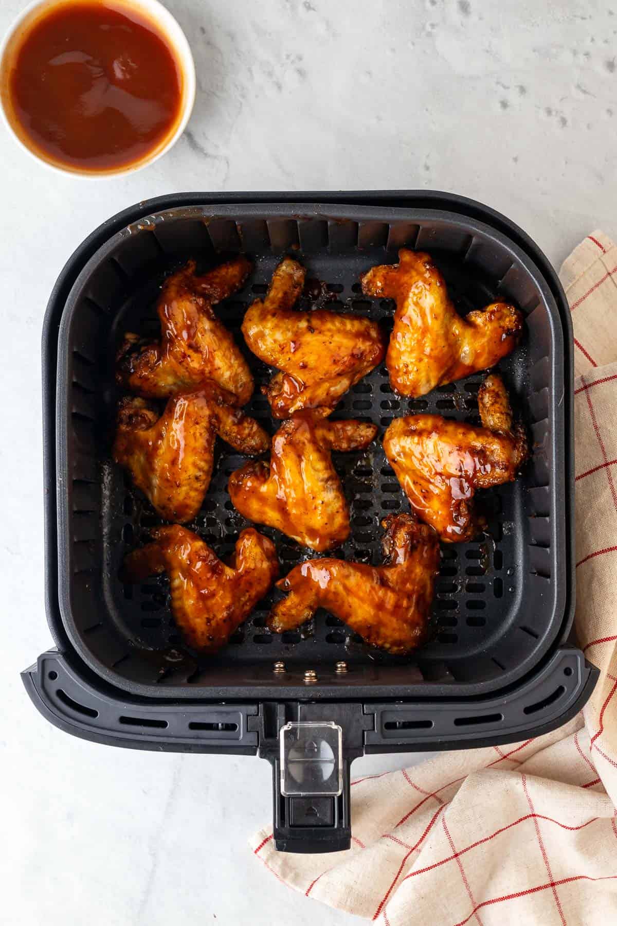 bbq sauce glazed chicken wings in the air fryer basket with the bbq sauce in a dish to one side and a decorative cloth on the other side