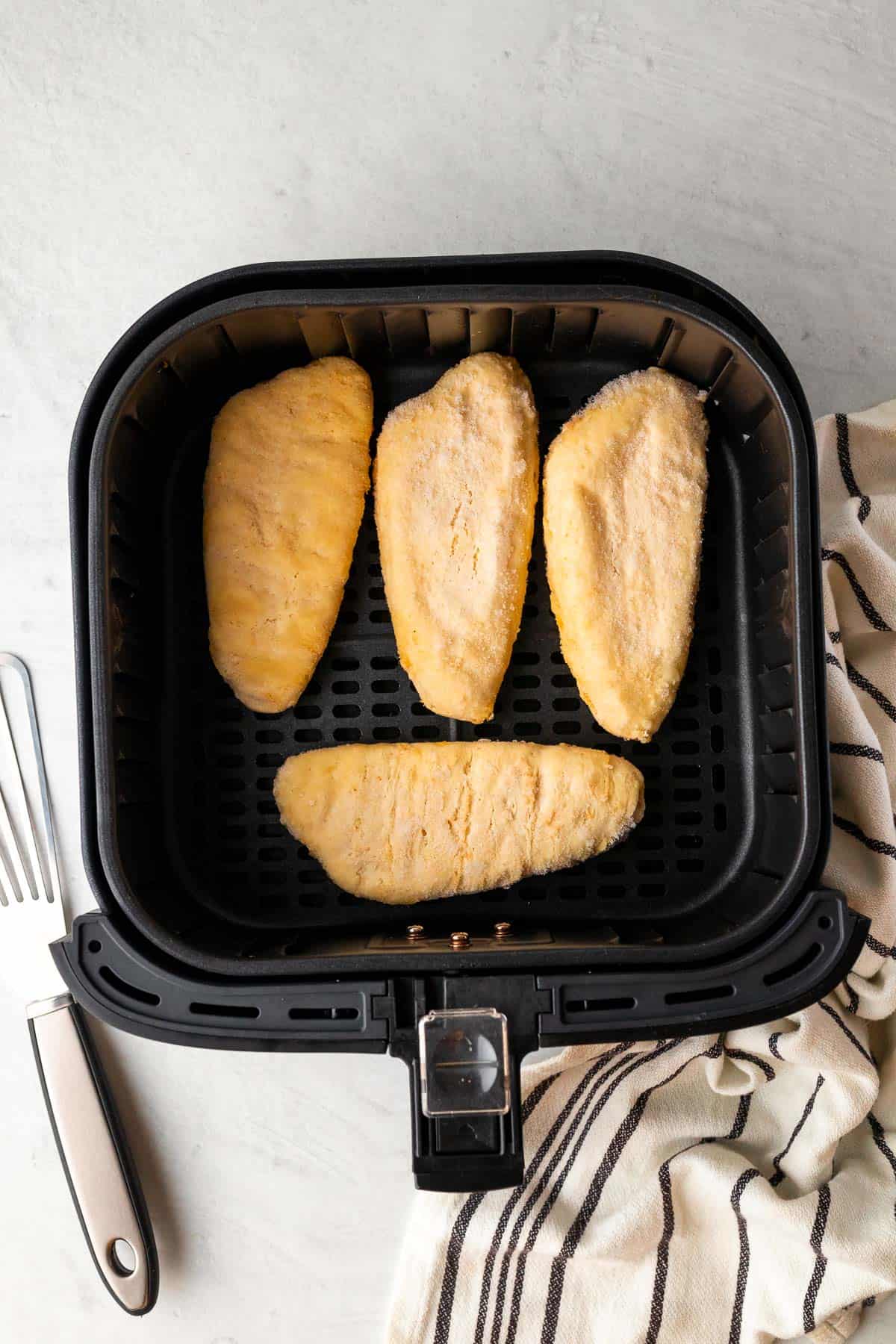 frozen fish fillets in the air fryer basket before cooking with a fish spatula and decorative cloth on either side of the air fryer basket