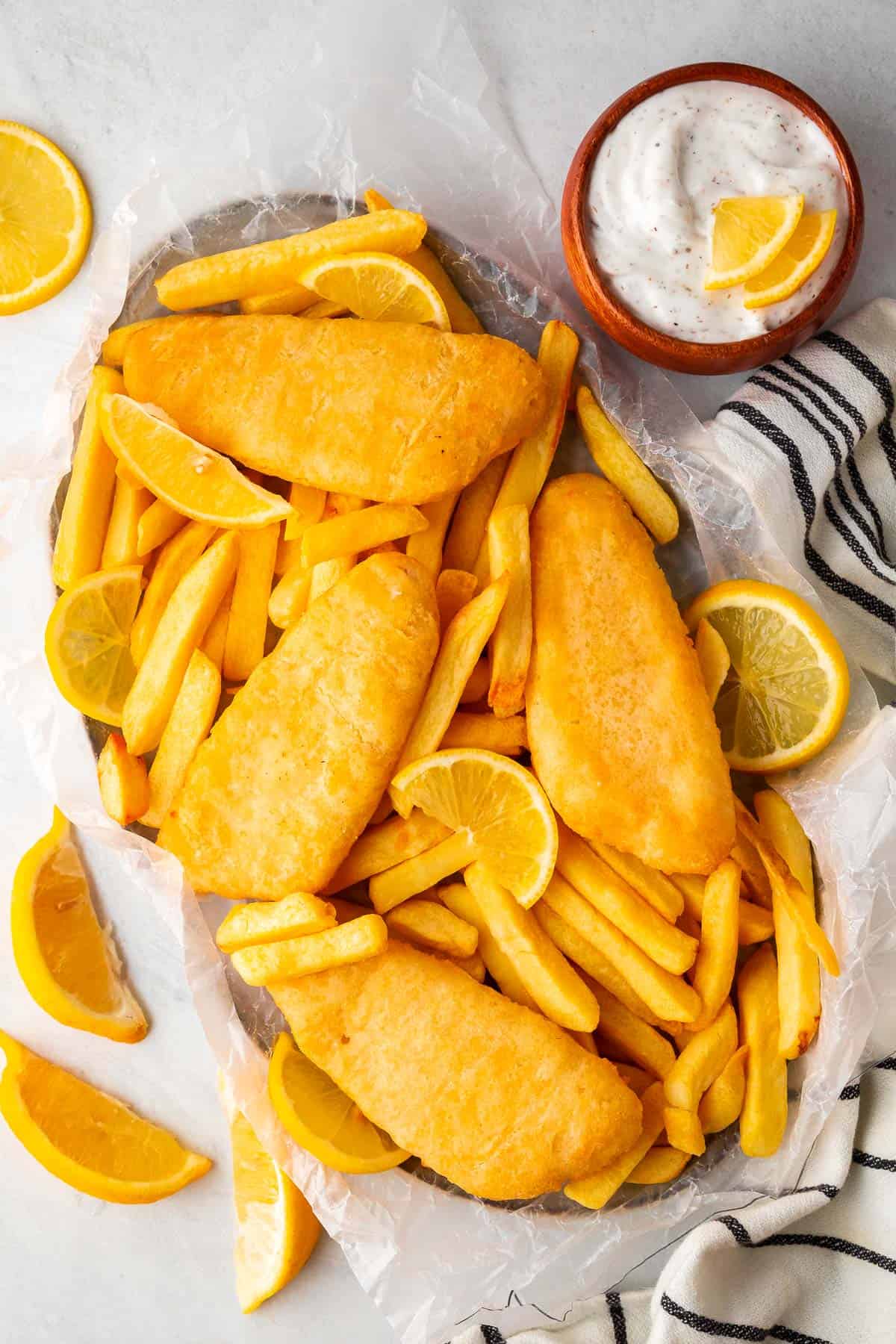 a serving platter full of fries, fish fillets and lemon slices and wedges. Tartar sauce and a decorative cloth to one side and more lemon wedges for decoration