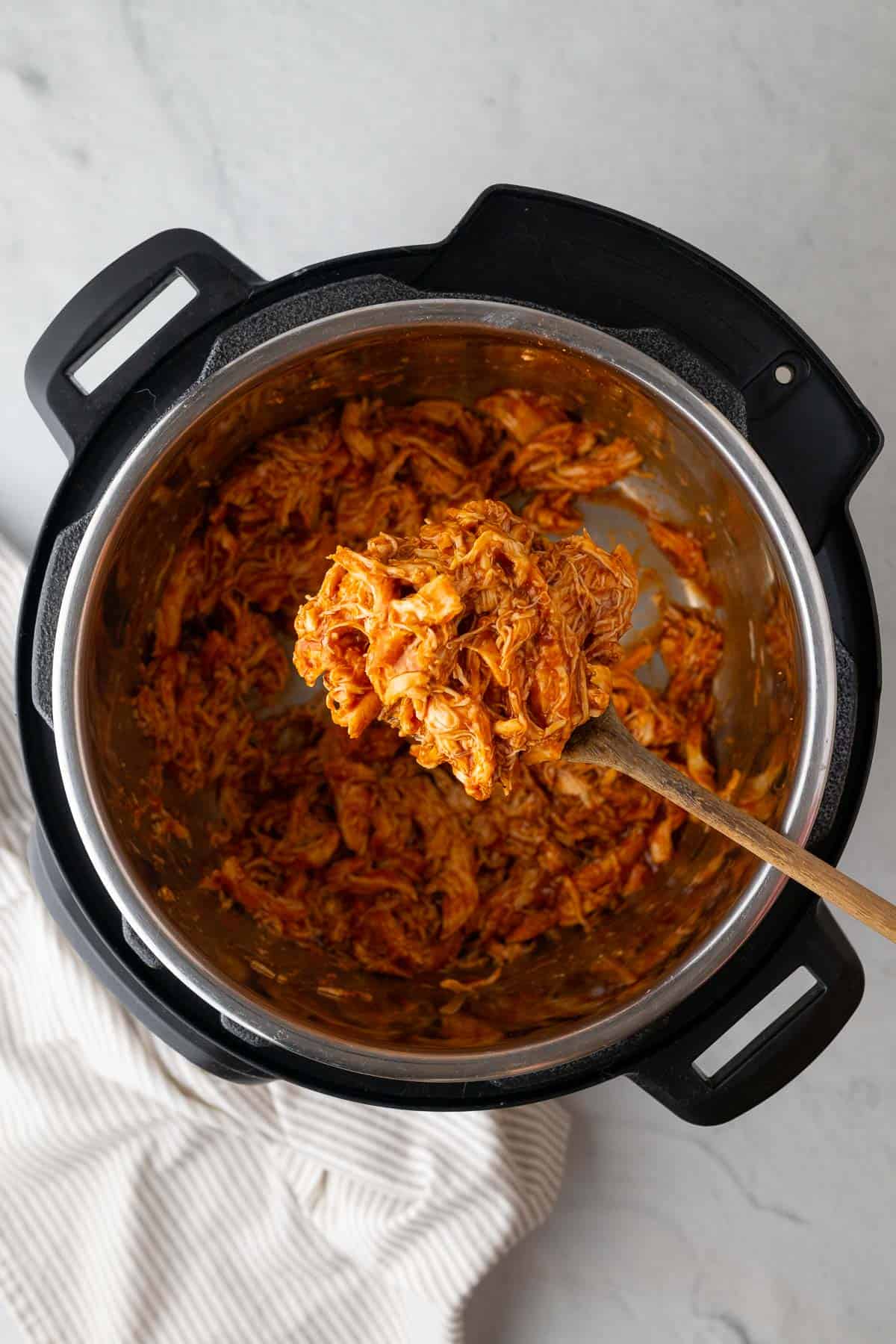 a wooden spoon holding up some bbq pulled chicken to the camera above the instant pot