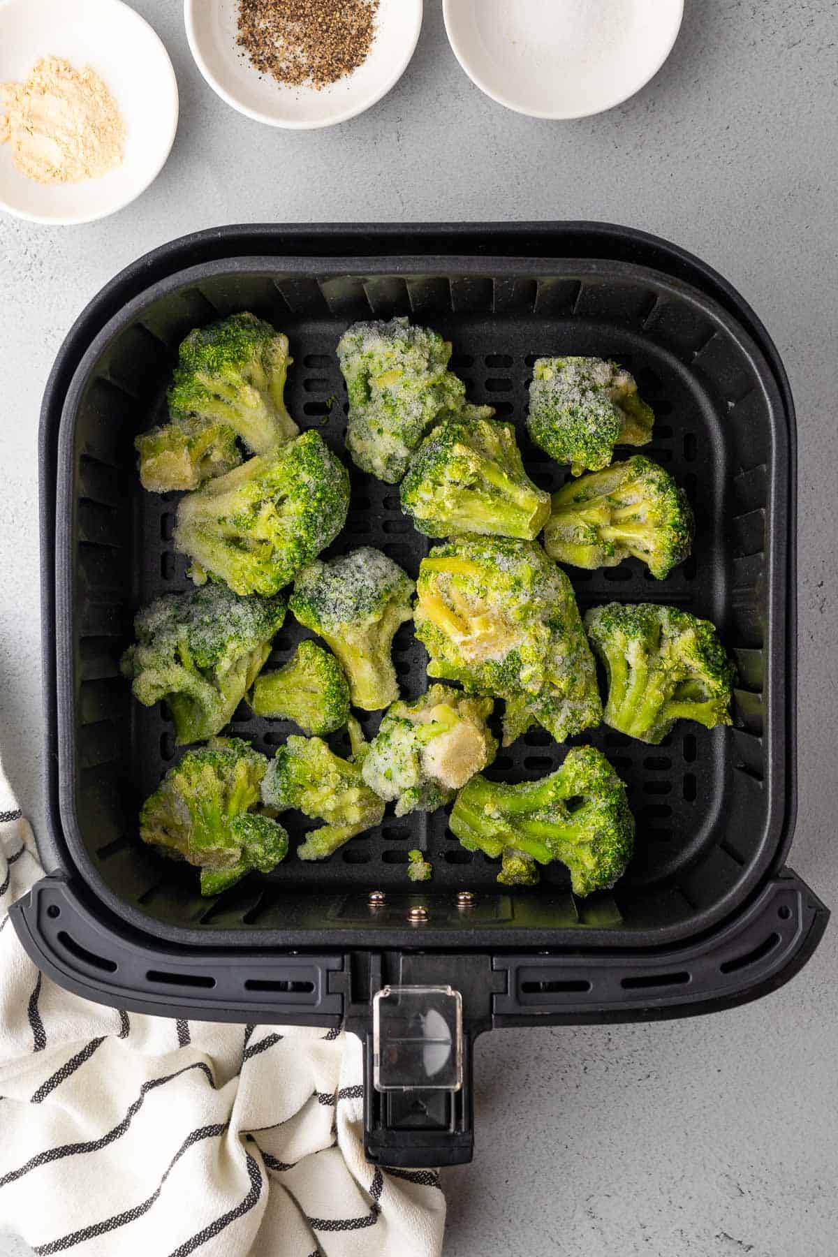 broccoli in the air fryer basket surrounded by the seasonings in little white dishes