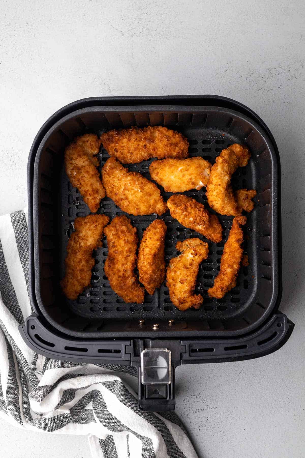 frozen chicken tenders in the air fryer basket after cooking with striped cloth to one side