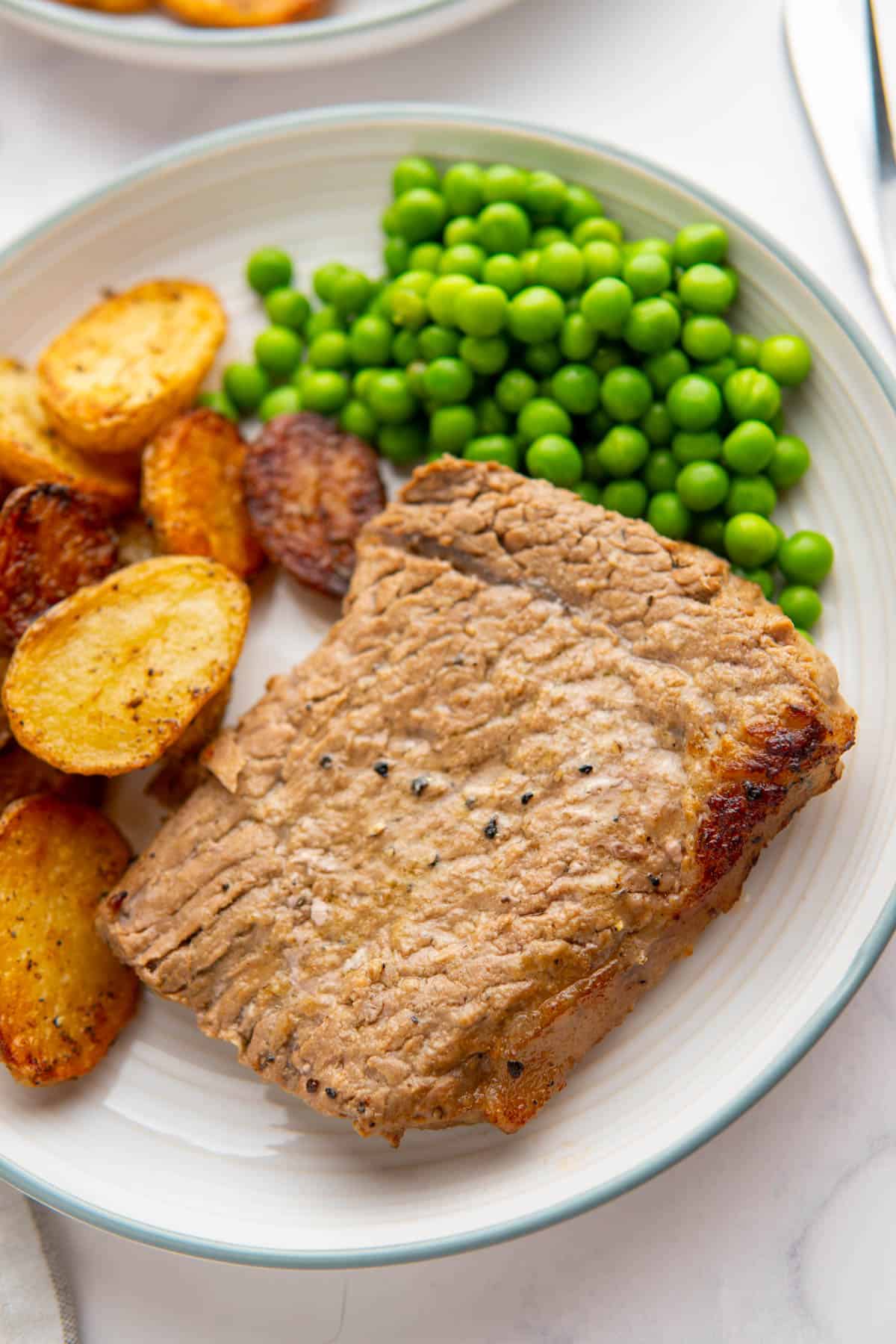 Close up on the marinated cube steak served on a plate with peas and roasted baby potatoes.