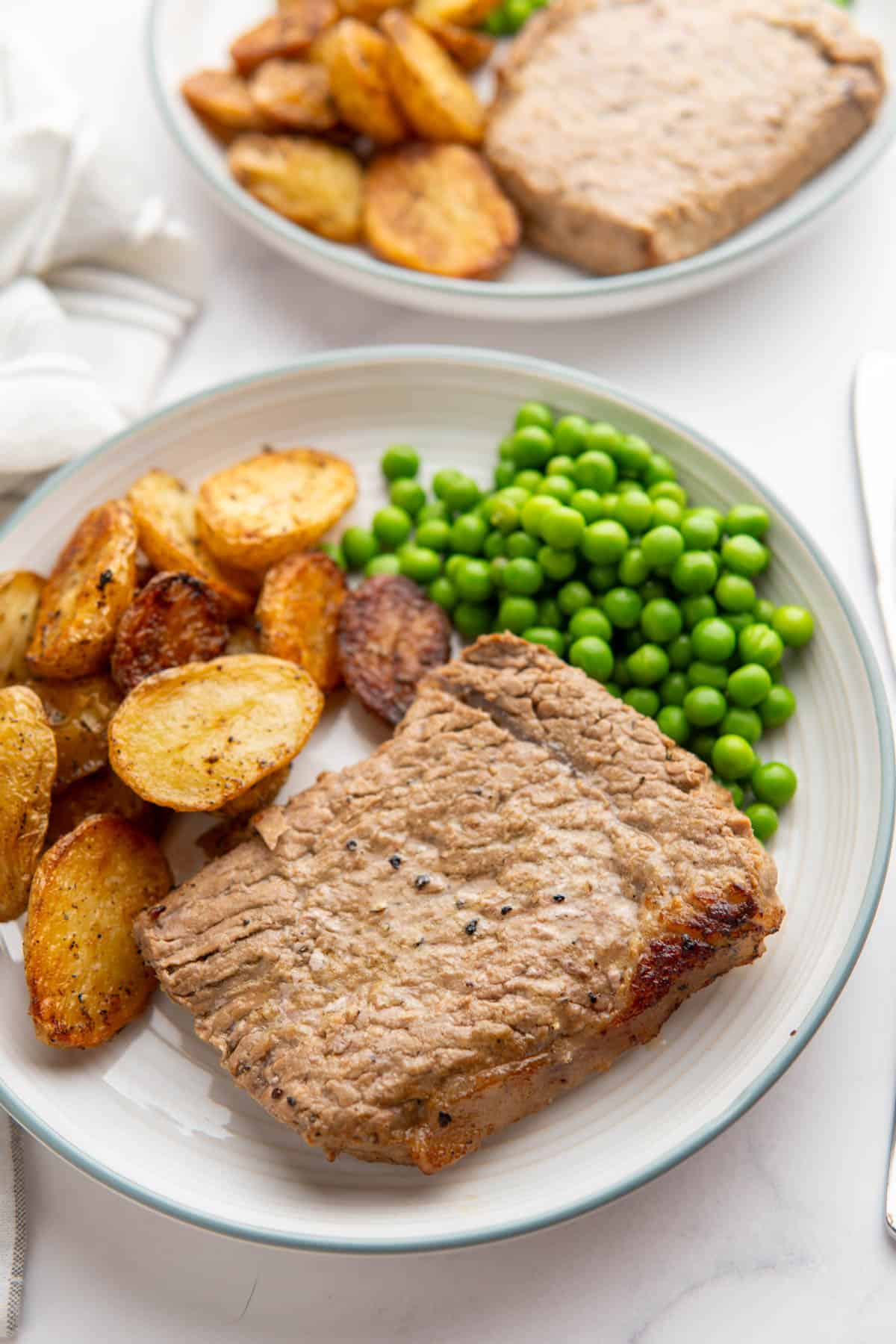 Marinated cube steak served on a plate with peas and roasted baby potatoes on a white background.