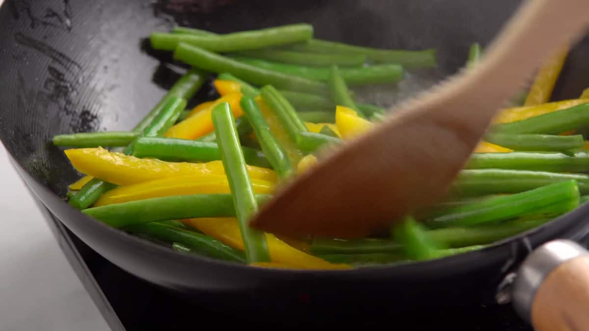 The green string beans and yellow bell pepper strips being stir fried in a large wok. 
