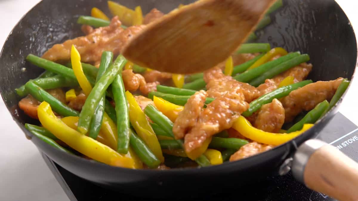 A wooden spoon mixing together the chicken, vegetables and sauce while they stir fry in a large wok. 