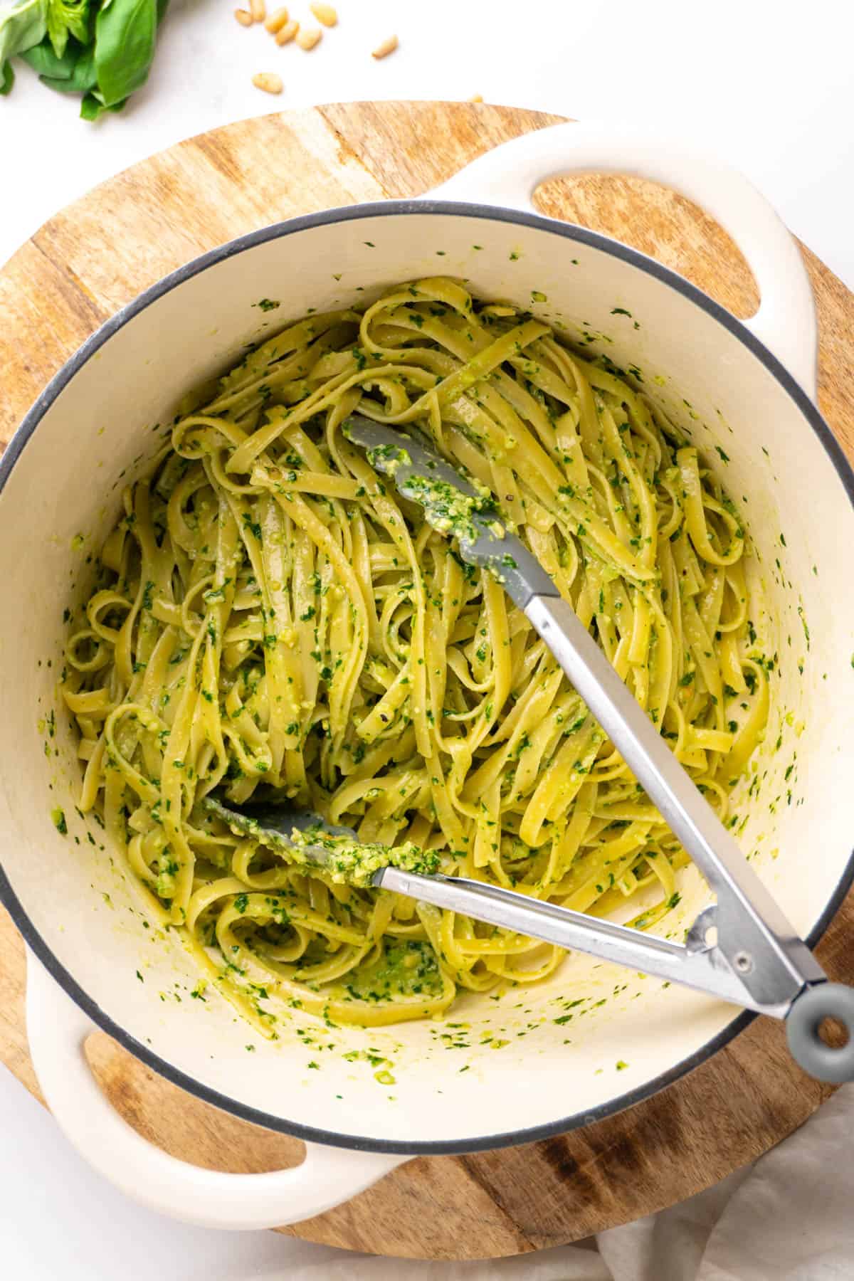 Cooked pasta and basil pesto tossed together in a large pot with a pair of tongs.