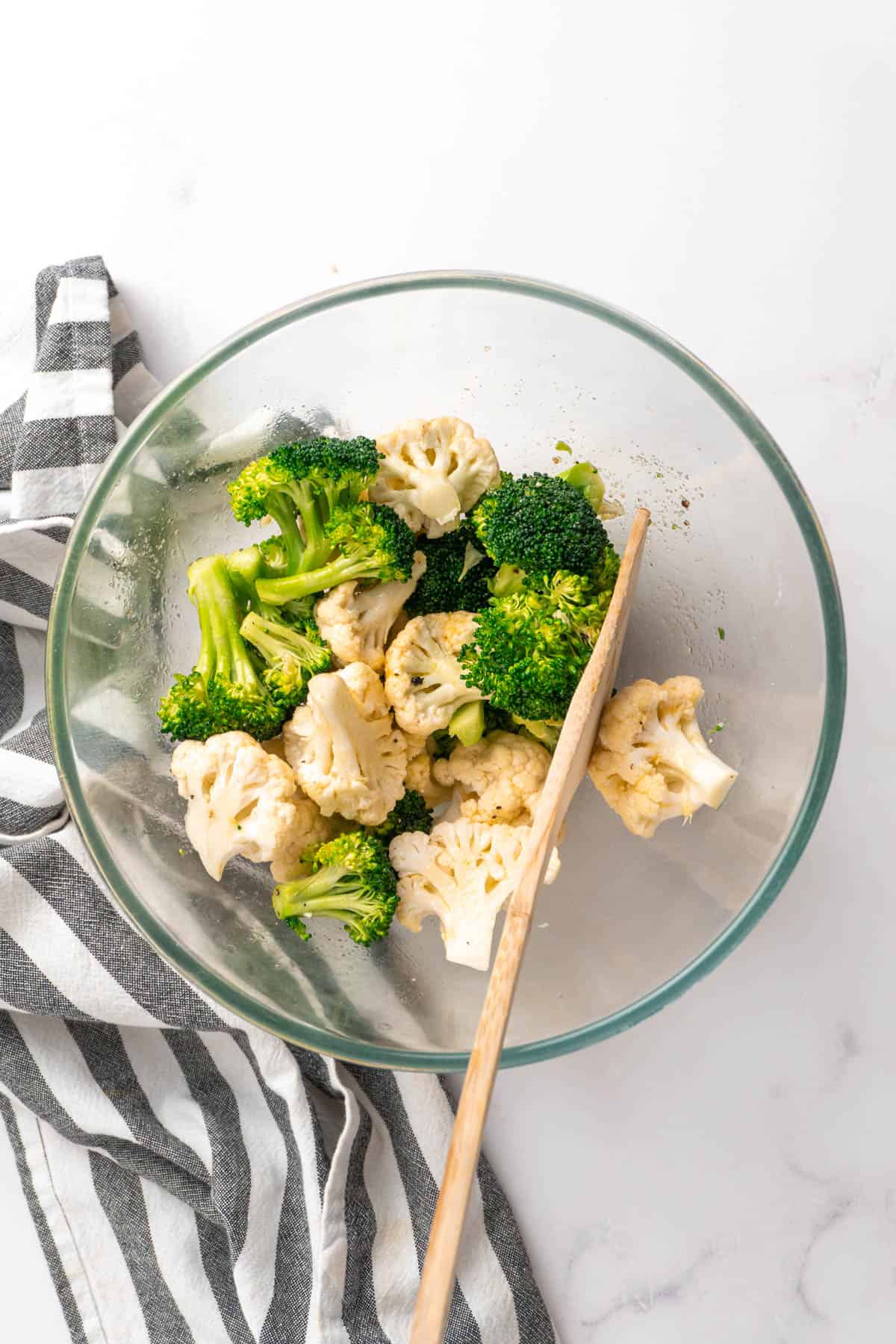 Cauliflower and broccoli are tossed with oil and seasonings in a large glass mixing bowl. 